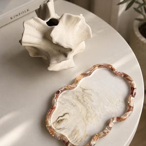 May include: A wavy-edged tray featuring a marbled white and beige center with a border of multicolored brown and tan resin chips. Beside it sits an off-white, sculptural ceramic vase with organic, folded, and petal-like edges.