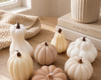 May include: A collection of seven pumpkin-shaped candles in various shades of white, cream, and tan. Each candle features a ribbed texture and a small brown stem at the top. The candles are arranged in a group, with some taller and some flatter.