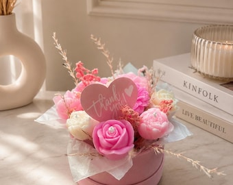 May include: A pink floral arrangement in a round box. The arrangement includes pink and white rose-shaped candles, pink flowers, and a heart-shaped card that says "Thank you". The box sits on a white surface.