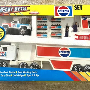 May include: A vintage Pepsi-branded toy set featuring a truck and trailer, a pickup truck, and other accessories. The set is packaged in a blue and yellow box with the words "Heavy Metal" and "Pepsi" prominently displayed. The set includes "Safe Non-Toxic Finish" and "Real Working Parts".