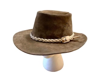 May include: A brown suede hat with a wide brim and a braided band. The hat has a slightly indented crown and a leather band with a decorative clasp. The hat is displayed on a white mannequin head.