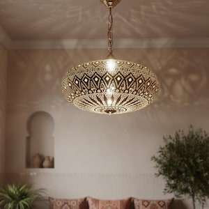 May include: A gold-colored, ornate pendant light fixture with an intricate, geometric pattern. The light hangs from a chain and casts decorative shadows on the ceiling. The fixture has a round, flattened shape, and is likely made of metal.