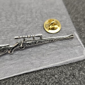 May include: A silver-toned rifle pin with a scope and a wood-grain pattern. The pin is on a white mesh bag with a gold pin back.