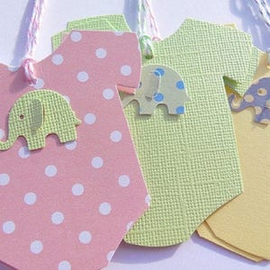 May include: Four baby onesie shaped gift tags with elephant designs. The tags are pink with white polka dots, green with a textured surface, yellow, and blue with white polka dots.