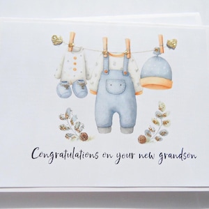 May include: A greeting card with a watercolor illustration of baby clothes hanging on a clothesline. The clothes include a onesie, a hat, a shirt, and booties. The text "Congratulations on your new grandson" is written in cursive.