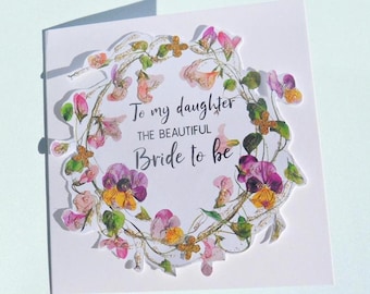Könnte beinhalten: Eine quadratische Grußkarte mit einem zentralen Blumenkranz aus rosa, violetten und gelben Stiefmütterchen mit goldenen Glitzerakzenten. Der Text in der Mitte lautet, To my daughter THE BEAUTIFUL Bride to be, in schwarzer Schrift.