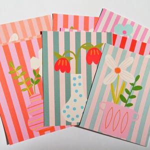May include: A selection of greeting cards with vertical striped patterns in pink, orange, and teal. Each card displays a floral illustration in a vase. The cards are arranged in a fan shape, offering a variety of designs.