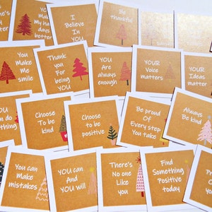 May include: A selection of square cards featuring motivational quotes and Christmas tree designs. The cards have a brown backdrop with white text and are arranged on a white surface. Messages include "You Make Me Smile" and "Choose to be kind."