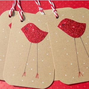 May include: Three kraft paper gift tags with red glitter bird designs and white polka dots. Each tag has a red and white twine string for hanging. The tags are on a red background.