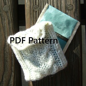 Puede incluir: Una bolsa de punto de ganchillo blanca con un patrón de punto de cable. La bolsa está sentada sobre una superficie de madera con un sobre azul y blanco debajo. El texto "PDF Pattern" está impreso en la bolsa.