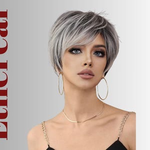 Puede incluir: Una mujer con una peluca corta y plateada. Lleva pendientes de aro dorados y un collar de oro. La palabra "Ethereal" está impresa verticalmente en rojo en el lado izquierdo de la imagen. La mujer tiene ojos azules y está maquillada.