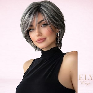 May include: A woman wearing a short, layered wig with a mix of gray and white strands. The wig has a side-swept fringe and is styled to frame the face. She is wearing a black turtleneck top. The background is a soft pink.