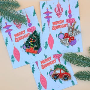 Può includere: Tre biglietti d'auguri natalizi con la scritta "MERRY & BRIGHT" e decorazioni di perline. I biglietti presentano un albero di Natale, una stella e un'auto rossa che trasporta un albero di Natale. Lo sfondo ha ornamenti rosa e viola.