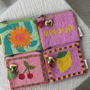 Könnte beinhalten: Vier Perlen-Münzbörsen mit Reißverschlüssen. Die Geldbörsen haben farbenfrohe Designs: eine Sonne, das Wort "AMOUR", Kirschen und eine Zitrone. Jede Geldbörse hat einen goldenen Herzanhänger und eine Quaste.