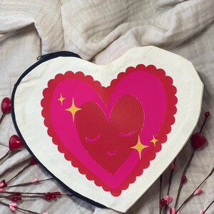 Op de afbeelding: Een witte hartvormige canvas tas met een roze en rood hartontwerp. Het hart heeft een glimlachend gezicht met gesloten ogen en gouden glitteraccenten.