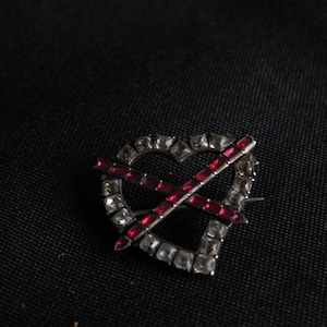 May include: A silver heart-shaped brooch with a red rhinestone cross design.