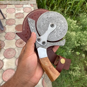 Może przedstawiać: Srebrny nóż do pizzy z drewnianym uchwytem i skórzaną pochwą. Nóż ma wzorzystą klingę i okrągłe koło tnące.