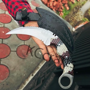 May include: A black leather sheath with a silver karambit knife with a honeycomb pattern handle. The knife has a ring on the end of the handle.