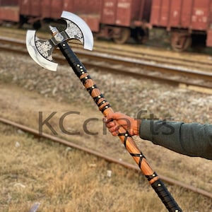 Puede incluir: Un hacha de doble hoja con cabeza plateada y mango de madera. La cabeza del hacha presenta tallas ornamentadas. El mango está envuelto en cuero negro y adornado con tachuelas de latón. El texto "KC Crafts" es visible.