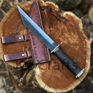 May include: A fixed-blade knife with a dark, textured metal blade and a black handle, featuring a silver bolster and pommel. It is accompanied by a brown leather sheath with decorative stitching and two leather belt loops with silver rivets.
