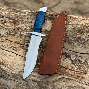 Puede incluir: Un cuchillo de caza artesanal con una hoja de acero pulido y un mango azul y plateado. El cuchillo va acompa&ntilde;ado de una funda de cuero marr&oacute;n. El cuchillo est&aacute; sobre un trozo de madera.