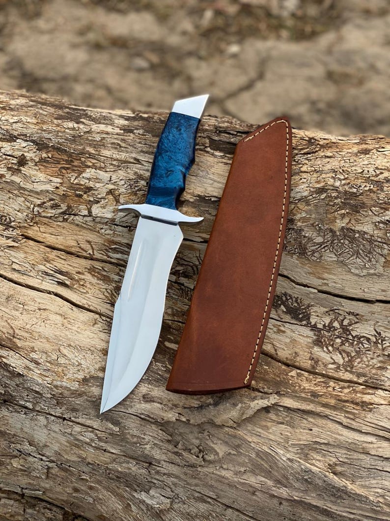 Puede incluir: Un cuchillo de caza artesanal con una hoja de acero pulido y un mango azul y plateado. El cuchillo va acompa&ntilde;ado de una funda de cuero marr&oacute;n. El cuchillo est&aacute; sobre un trozo de madera.
