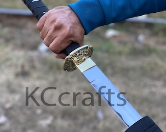Puede incluir: Una espada katana con empuñadura negra y guarda dorada. La hoja es plateada y refleja la luz. El texto "KCcrafts" está impreso en la hoja. La espada es sostenida por una persona que lleva una camisa azul de manga larga.