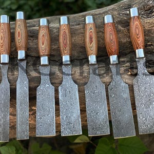 Op de afbeelding: Set van acht beitels met houten handvatten en een gehamerd stalen blad. De beitels hebben een houten handvat met een rood en bruin houtnerfpatroon. De bladen zijn gemaakt van gehamerd staal met een patroon van swirls en lijnen.