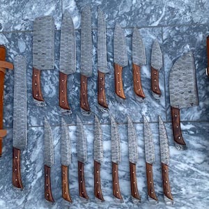 May include: A collection of various kitchen knives with wooden handles and patterned blades, arranged on a marble surface. Two brown leather knife rolls with buckle closures are on either side of the knives.