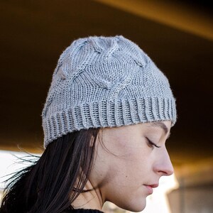 Side profile of a young woman wearing a light gray knit Seagulls Hat and a black sweater, standing under a bridge. This is an easy beanie knitting pattern.