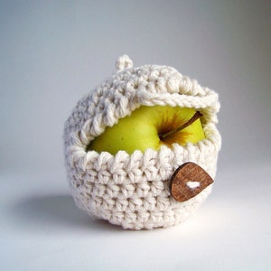 A green apple is wrapped in an off-white crochet apple cozy with a small brown button closure on a white background.