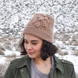 Puede incluir: Una persona lleva un gorro de punto marrón con un patrón de punto de cable y una chaqueta verde. El gorro está tirado sobre las orejas y la chaqueta está cerrada con cremallera.