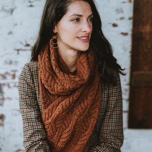 May include: A woman wearing a brown cable knit scarf with a plaid jacket. She is wearing gold hoop earrings with a brown leather accent.