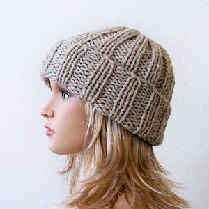 May include: A beige knitted beanie hat with a ribbed texture. The hat is made with a simple stitch pattern and has a slouchy fit.