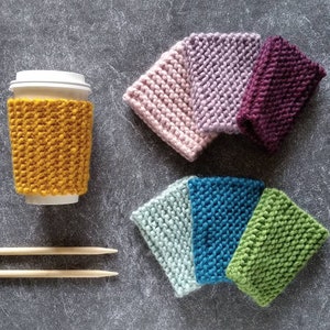 May include: A set of six knitted cup cosies in various colours, including mustard yellow, pink, purple, teal, green, and light blue. The cosies are made with a chunky yarn and have a simple, textured stitch.