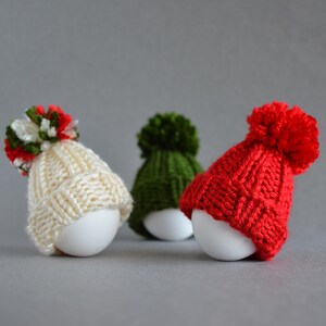 May include: Three white eggs each wearing a knitted hat with a pom-pom. One hat is cream-colored with a red, green, and white pom-pom, one is green, and the other is red. The hats are miniature and appear handmade.