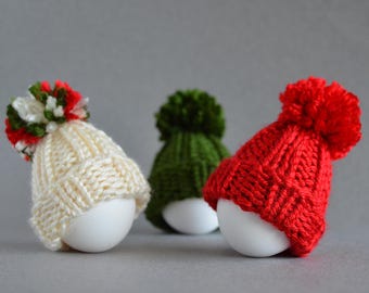 May include: Three white eggs each wearing a knitted hat with a pom-pom. One hat is cream-colored with a red, green, and white pom-pom, one is green, and the other is red. The hats are miniature and appear handmade.