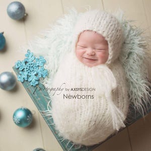 Op de afbeelding: Een pasgeboren baby die een witte gebreide muts en een witte gebreide wikkel draagt, lachend. De baby ligt op een blauw en wit pluche deken. De afbeelding bevat de tekst "Photography by AXSYSDESIGN Newborns".