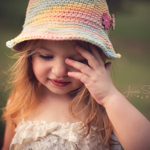 A cute girl with ginger hair wearing a pastel-colored crochet sun hat adorned with two buttons and a creamy ruffled dress, standing in an outdoor setting with a blurred green backdrop.