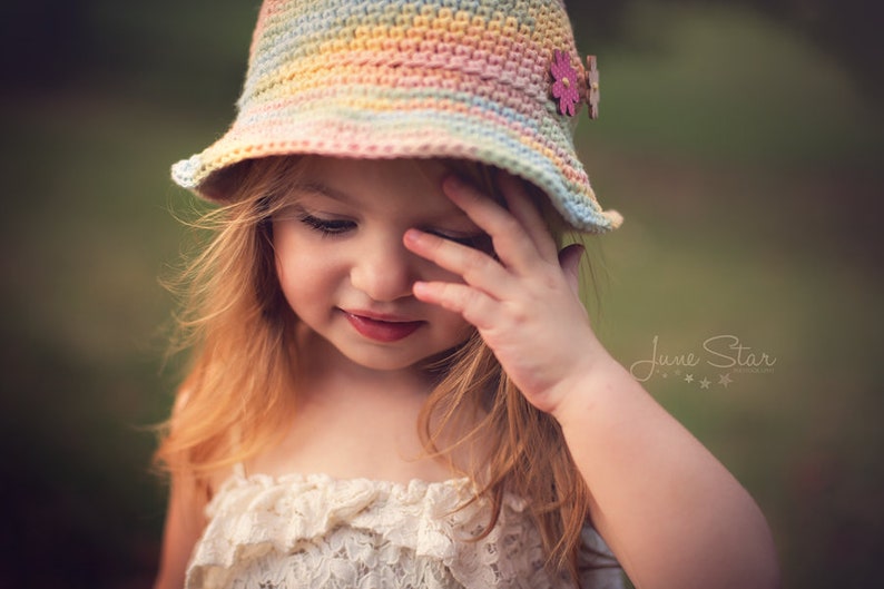 A cute girl with ginger hair wearing a pastel-colored crochet sun hat adorned with two buttons and a creamy ruffled dress, standing in an outdoor setting with a blurred green backdrop.