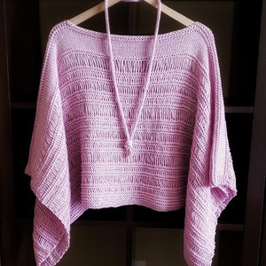 Light pink knitted sweater with wide sleeves and boatneck neckline, an a knit i-cord necklace displayed on a wooden hanger in a dark, open wardrobe.