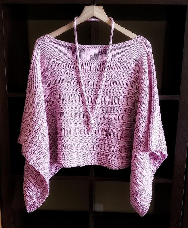 Light pink knitted sweater with wide sleeves and boatneck neckline, an a knit i-cord necklace displayed on a wooden hanger in a dark, open wardrobe.