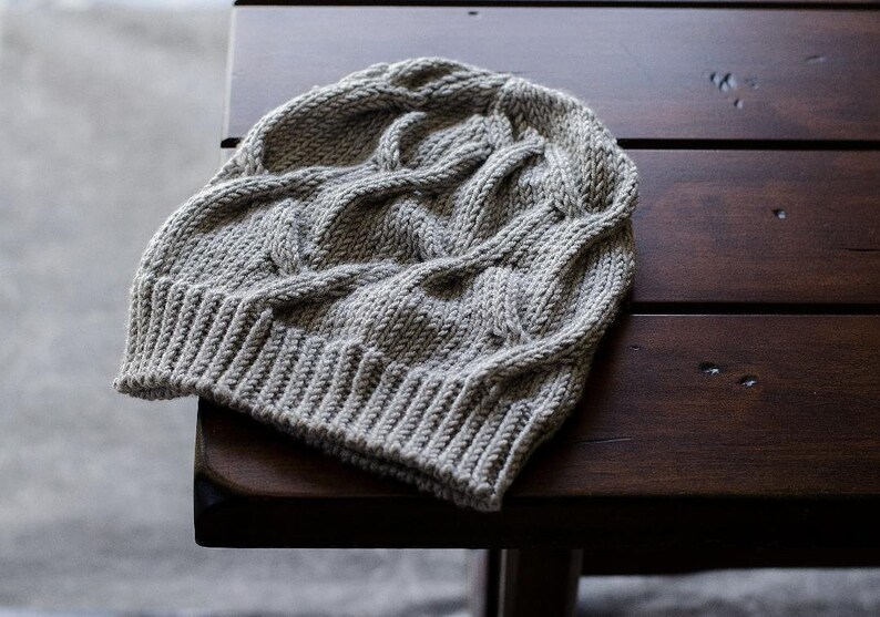A light gray knit Seagulls Hat with a ribbed brim and cable stitch pattern laid flat on a dark brown wooden table. it is an easy beanie knitting pattern.
