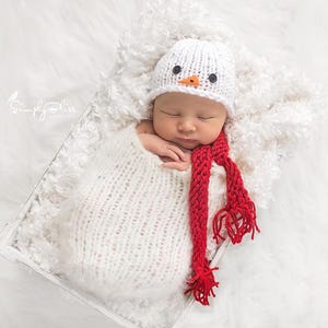 Op de afbeelding: Een pasgeboren baby gewikkeld in een witte gebreide deken en met een witte sneeuwmanhoed met zwarte knoopogen en een oranje wortelneus. Een rode gebreide sjaal is om de nek van de baby gedrapeerd.
