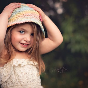 A cute young girl holding onto a pastel color crochet sun hat on her head, wearing a lacy cream dress, with a soft focus background of green foliage.