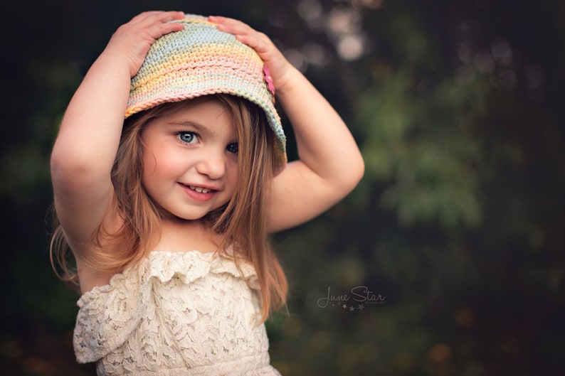 A cute young girl holding onto a pastel color crochet sun hat on her head, wearing a lacy cream dress, with a soft focus background of green foliage.