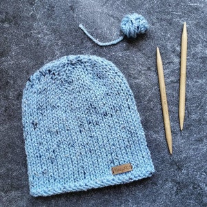 May include: A light blue knitted hat with a brown leather tag that says "Made by KC". The hat is sitting on a gray surface with two wooden knitting needles and a ball of blue yarn.