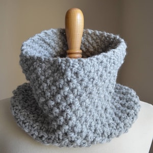 May include: A gray crocheted cowl neck warmer on a wooden display stand.