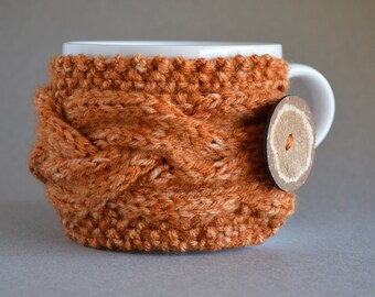 Può includere: Una tazza in ceramica bianca con un rivestimento in maglia arancione e bianco. Il rivestimento ha un design intrecciato e un bottone marrone.