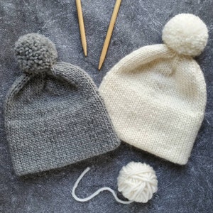 Puede incluir: Dos gorros de punto con pompones, uno gris y otro blanco, con agujas de tejer y una bola de lana.